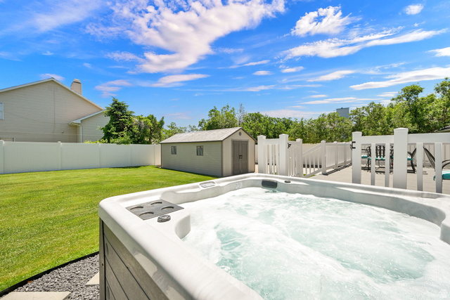 4792 Glasmann Way South Ogden, UT 84403 - Photo 23 of 70 Outdoor hot tub with textured gray siding and stone surround, accompanied by a spacious green lawn, a white privacy fence, an outdoor storage sheds, and a composite deck with white railings