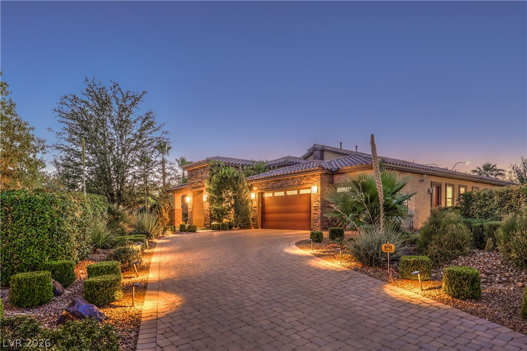 7119 Arabian Ridge Street Las Vegas, NV 89131 - Photo 4 of 73 An inviting brick paver driveway leads to a warm, well-lit entrance and garage, set against a backdrop of mature desert landscaping.