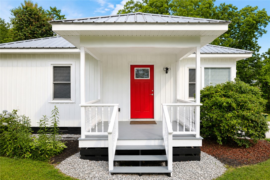605 Hampton Street Iva, SC 29655 - Photo 3 of 16 A vibrant red entry door beckons under a covered porch, set against clean white siding and lush greenery.