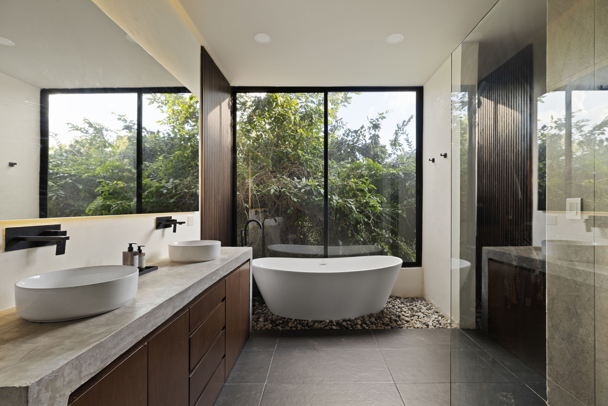 6 Country Club, Unit 4 Nome, TX 77629 - Photo 21 of 45 This spa-like primary bathroom delivers a masterclass in modern biophilic design, offering a serene oasis centered around panoramic views of the lush tropical canopy. The luxurious space highlights a freestanding soaking tub resting on a decorative bed of polished river rocks beneath an immense picture window, while a spacious walk-in shower and dual white vessel sinks sit atop a custom-poured concrete countertop framed by gorgeous vertical wood-slat accent walls.