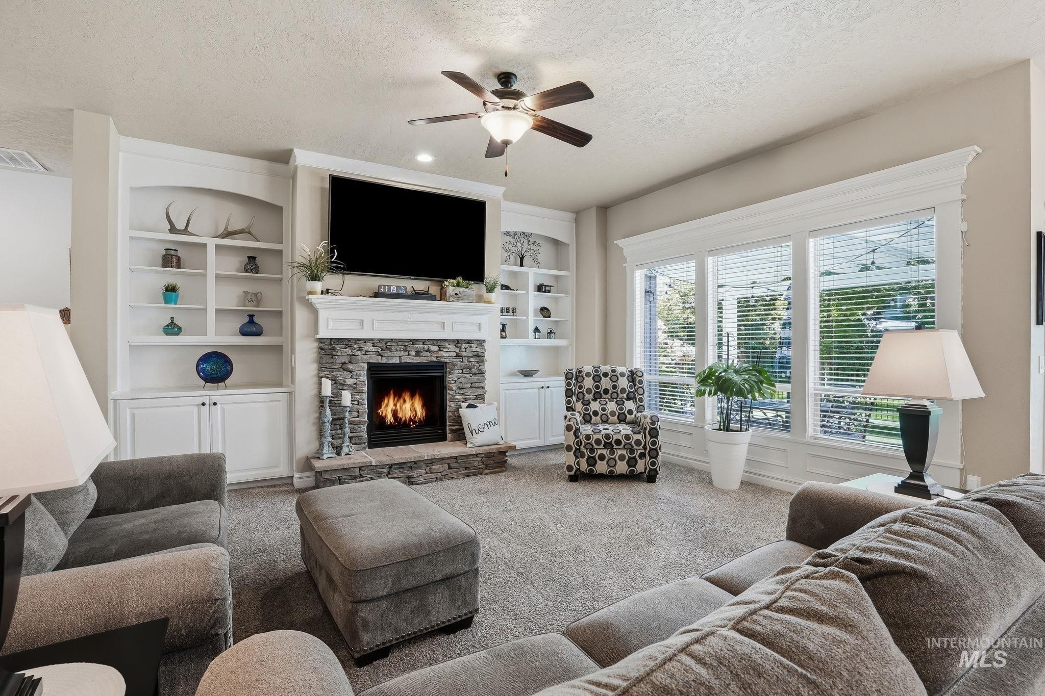 3969 North Hollymount Way Meridian, ID 83646 - Photo 7 of 47 Living area featuring a stone-faced fireplace with a white mantel, built-in shelving, a ceiling fan, and large windows with blinds