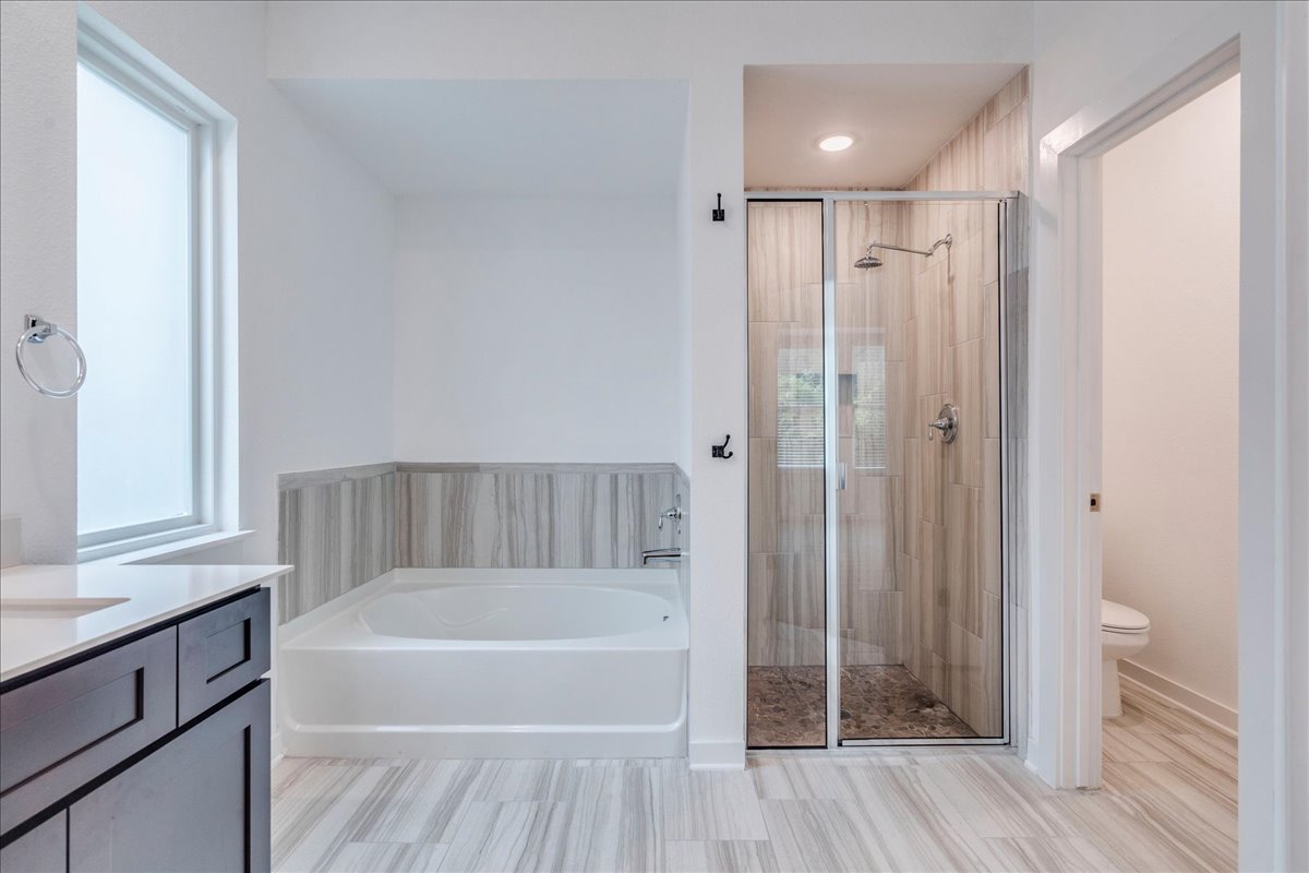 8008 Prairie Rye Drive Lago Vista, TX 78645 - Photo 8 of 40 Modern bathroom featuring a built-in soaking tub with tile surround, a glass-enclosed shower with tile walls and floor, wood-finish flooring, a dark wood-finish vanity with a light-colored countertop, and a frosted window