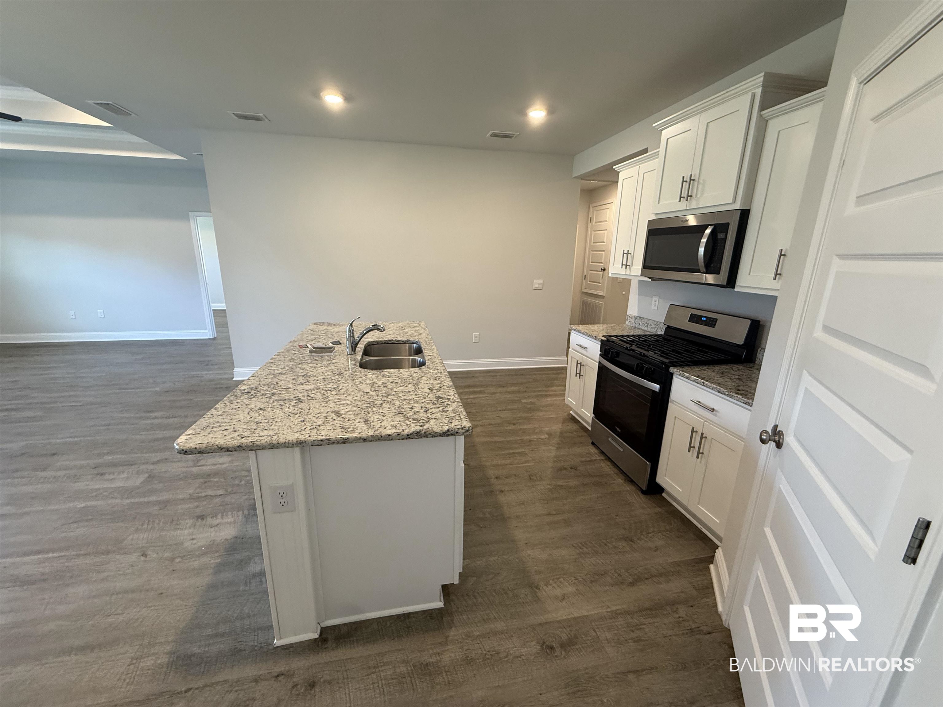 23247 Lieutenant Avenue Foley, AL 36535 - Photo 23 of 47 Open-concept kitchen featuring an island with a double basin sink, white cabinetry with silver hardware, stainless steel appliances, speckled countertops, and wood-finish flooring