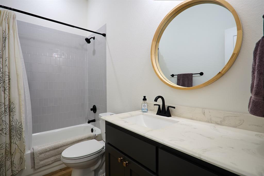 358 Glade Valley Road Azle, TX 76020 - Photo 19 of 35 Full bathroom featuring a dark vanity with a white countertop and integrated sink, a circular wood-frame mirror, matte black fixtures, and a white tiled shower-tub combination