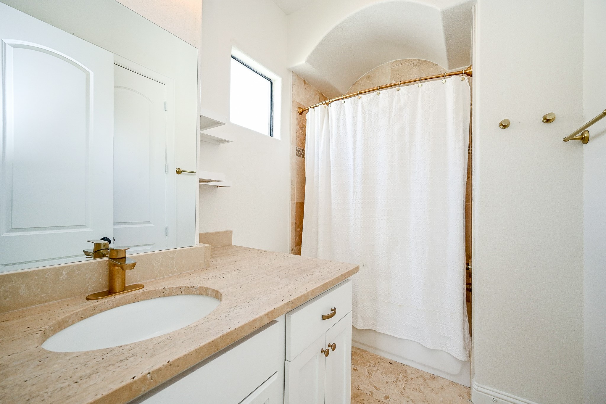 1825 West 24th Street Houston, TX 77008 - Photo 28 of 32 This bathroom features a clean, modern design with a light-colored stone countertop, stylish brass fixtures, and a white shower curtain. It includes built-in shelving and a large mirror, creating a bright and open feel.