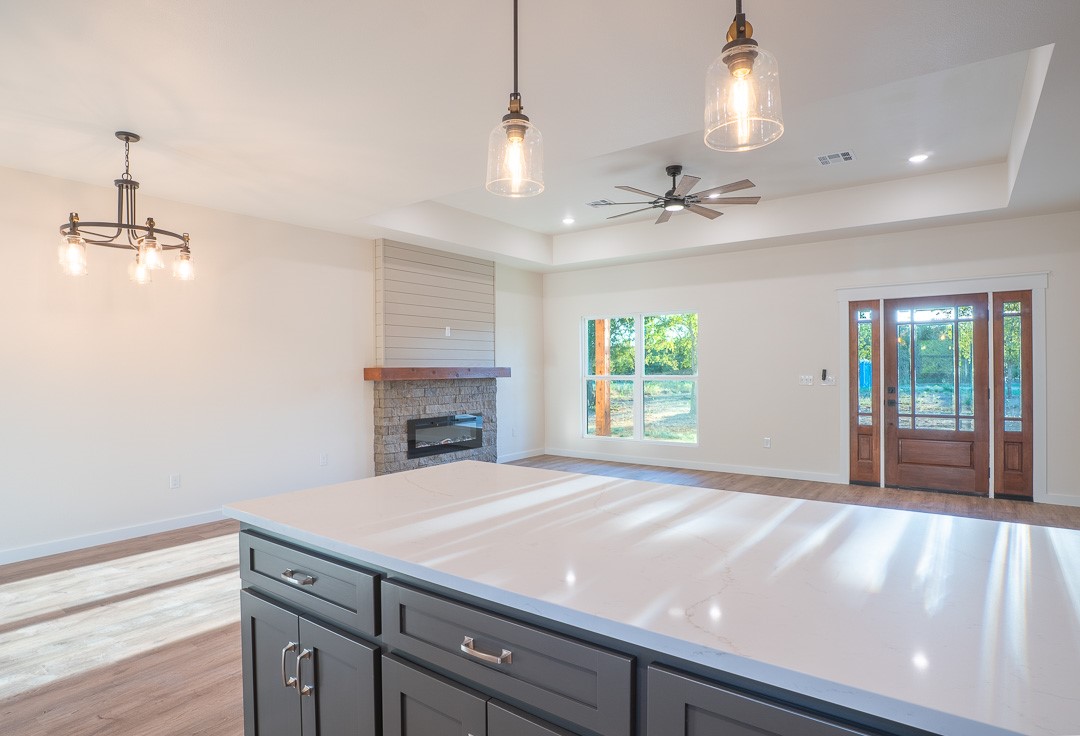 205 Blessings Way Colbert, OK 74733 - Photo 12 of 24 Kitchen island with dark cabinetry and a light countertop, wood-finish flooring, stone fireplace with a wood mantle, ceiling fan, and large windows