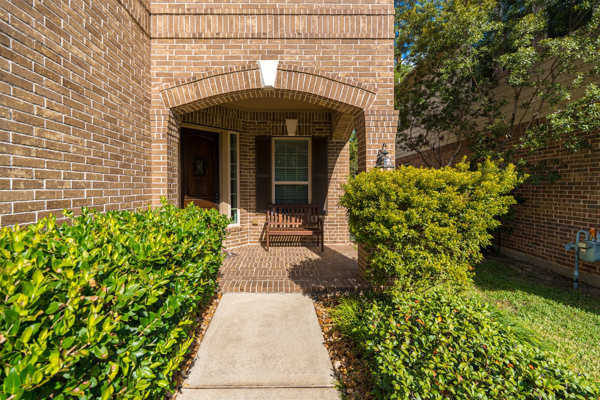 30 Fury Ranch Place The Woodlands, TX 77375 - Photo 9 of 47 Charming brick home entry with an inviting covered porch. Features a wooden door, a window with shutters, and a cozy bench. Flanked by manicured hedges and lush greenery, creating a welcoming atmosphere.