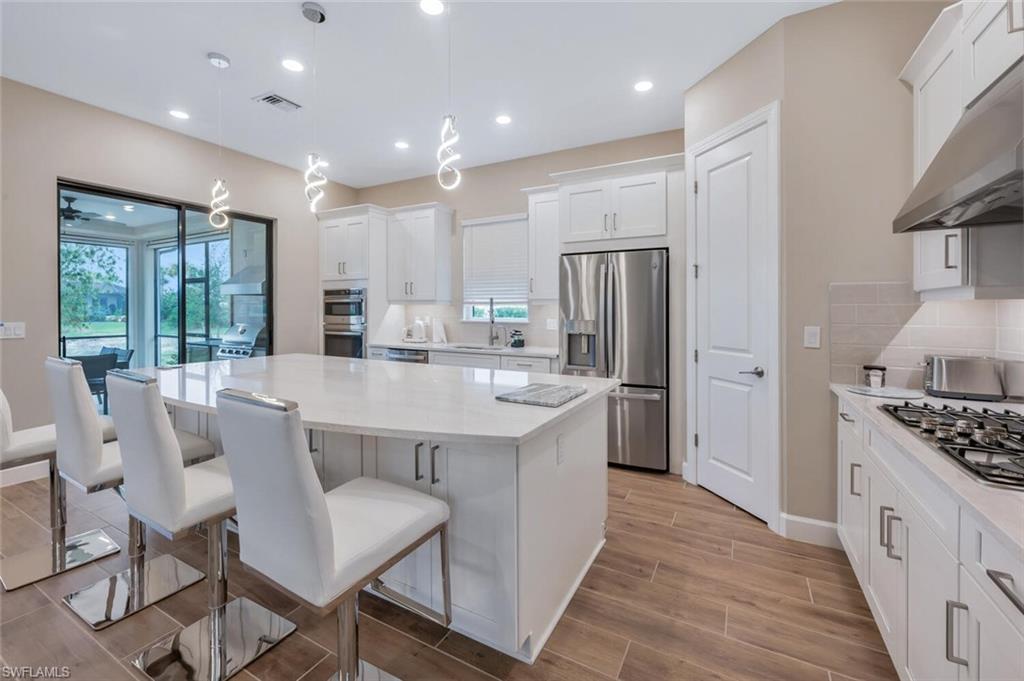8448 Rosa Court Naples, FL 34114 - Photo 11 of 24 Open-concept kitchen featuring an expansive central island with ample seating, white shaker-style cabinetry, wood-finish tile flooring, recessed lighting, and stainless steel appliances