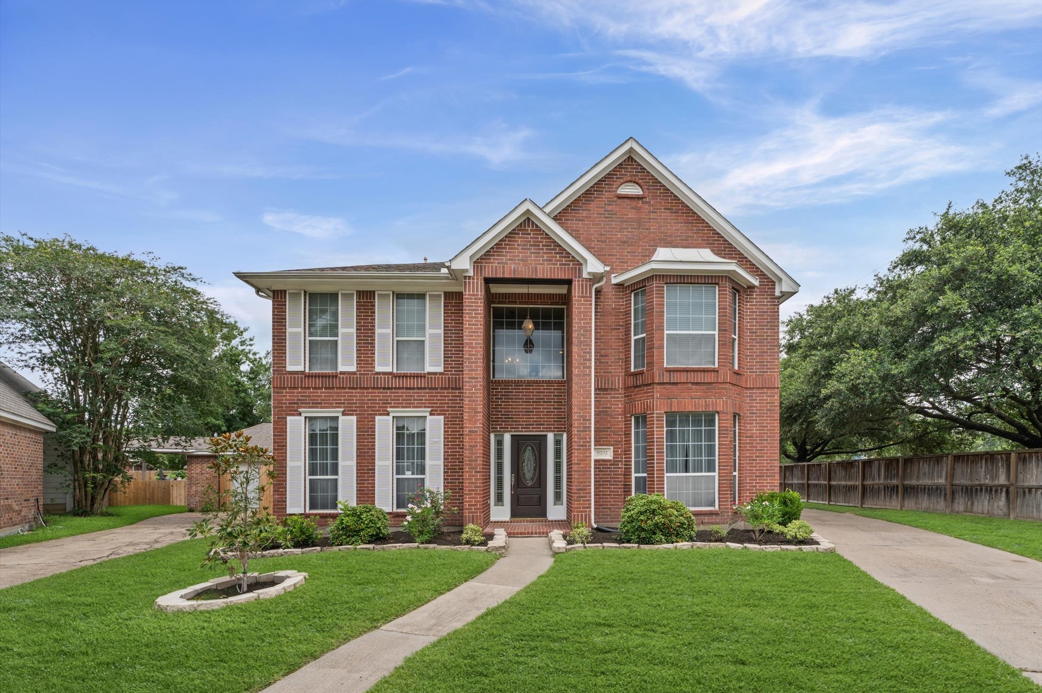9202 State Walk Circle Houston, TX 77064 - Photo 1 of 25 This is a two-story brick home with large windows and a well-maintained front yard. The symmetrical facade features a central entrance flanked by bay windows, and a driveway leading to a side garage. The house is framed by mature trees and a wooden fence, offering a classic suburban feel.