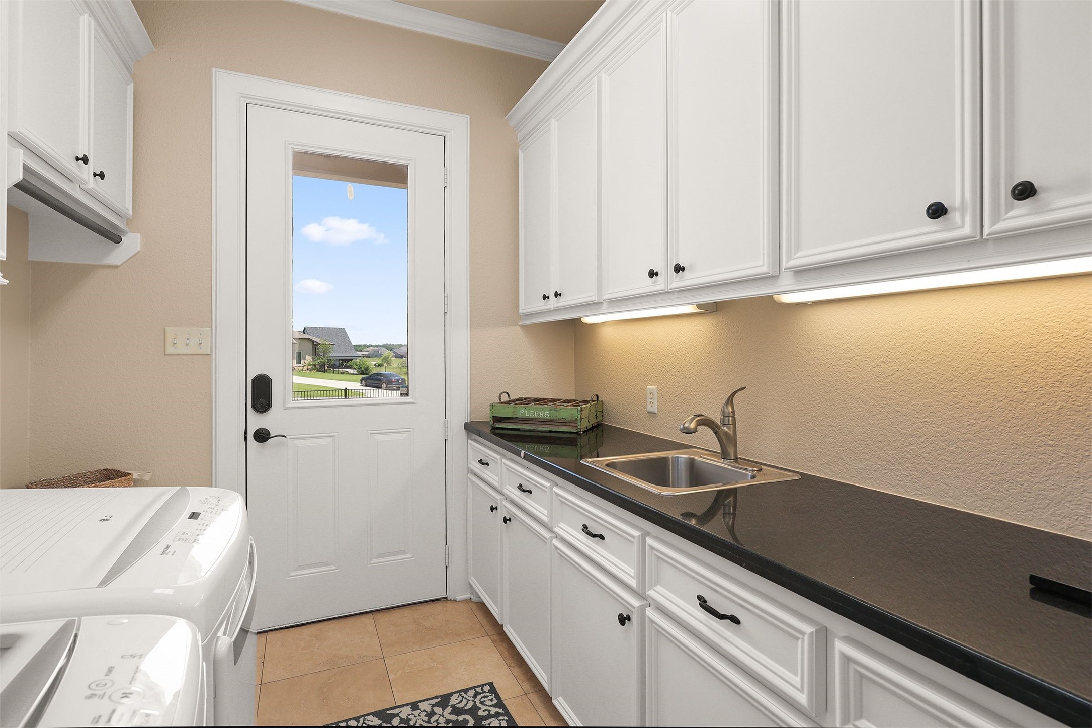 10896 Decatur Street Willis, TX 77318 - Photo 22 of 50 This bright and functional laundry room boasts ample white cabinetry, a sleek black countertop, a stainless steel sink, and a washer and dryer, all enhanced by natural light from a glass door leading outside.