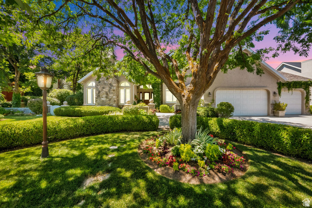 Manicured front yard with mature trees, stone veneer accents, arched windows, a two-car garage, and established landscaping