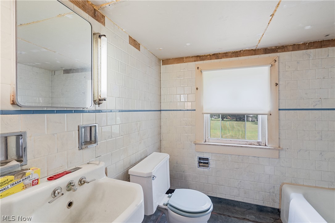 6324 Struthers Road Youngstown, OH 44514 - Photo 39 of 42 Bathroom featuring white tiled walls with a blue accent line, a built-in bathtub, a toilet, a sink with multiple faucets, and a window with a white roller shade