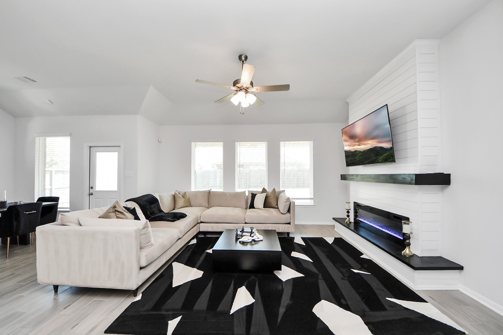 10603 Juniper Br Drive Rosharon, TX 77583 - Photo 23 of 50 This modern living room features a spacious L-shaped sofa, a sleek black and white rug, and a wall-mounted TV above an electric fireplace. Large windows provide ample natural light, while a ceiling fan offers comfort. The open layout flows into a dining area, creating an inviting space for relaxation and entertainment.