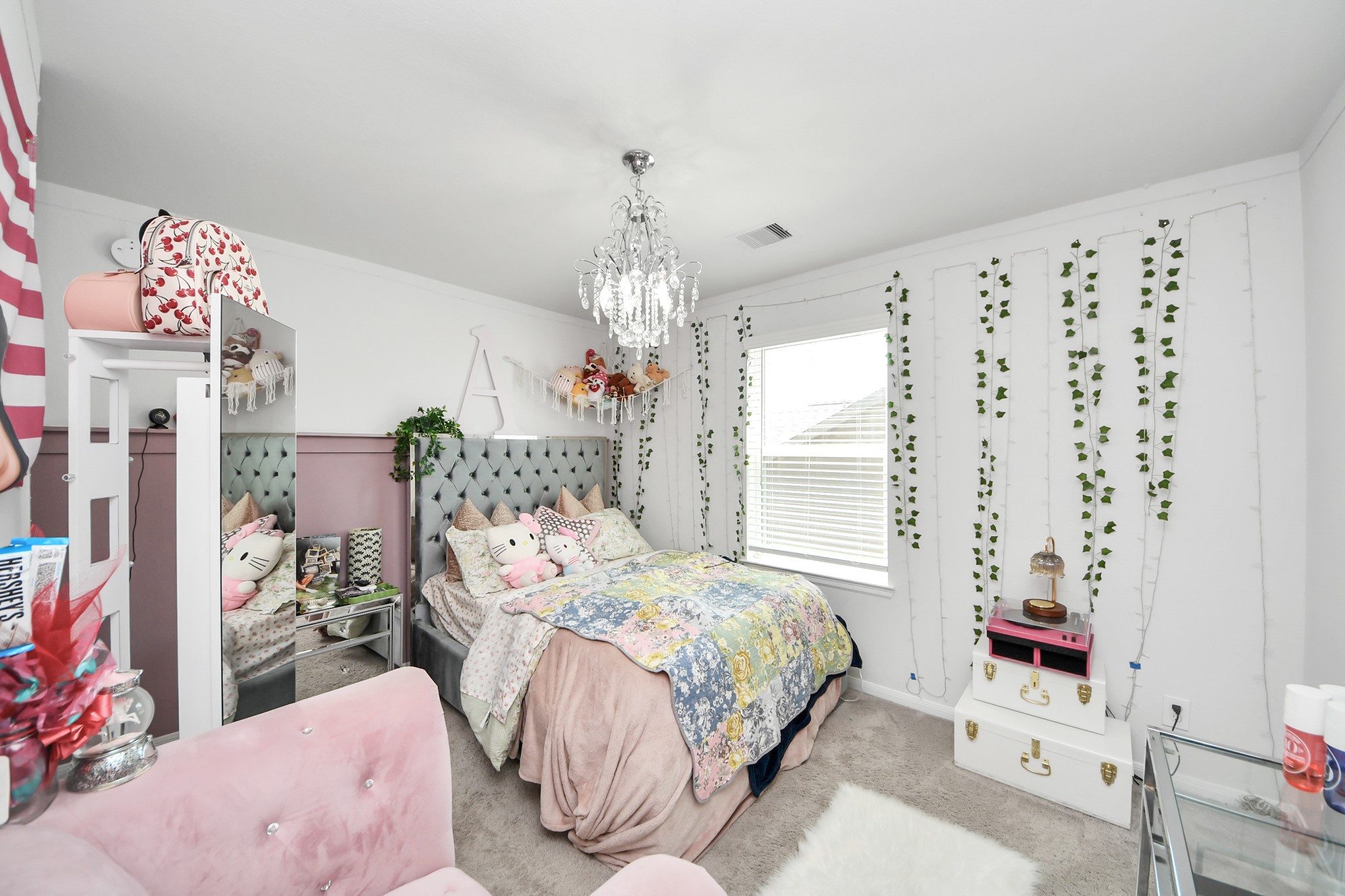 10603 Juniper Br Drive Rosharon, TX 77583 - Photo 34 of 50 This cozy bedroom features a plush, tufted headboard and a colorful, patterned bedspread. It has a modern chandelier, decorative wall vines, and ample natural light from a large window. The room is accented with a soft pink chair and stylish decor, creating a welcoming and vibrant atmosphere.