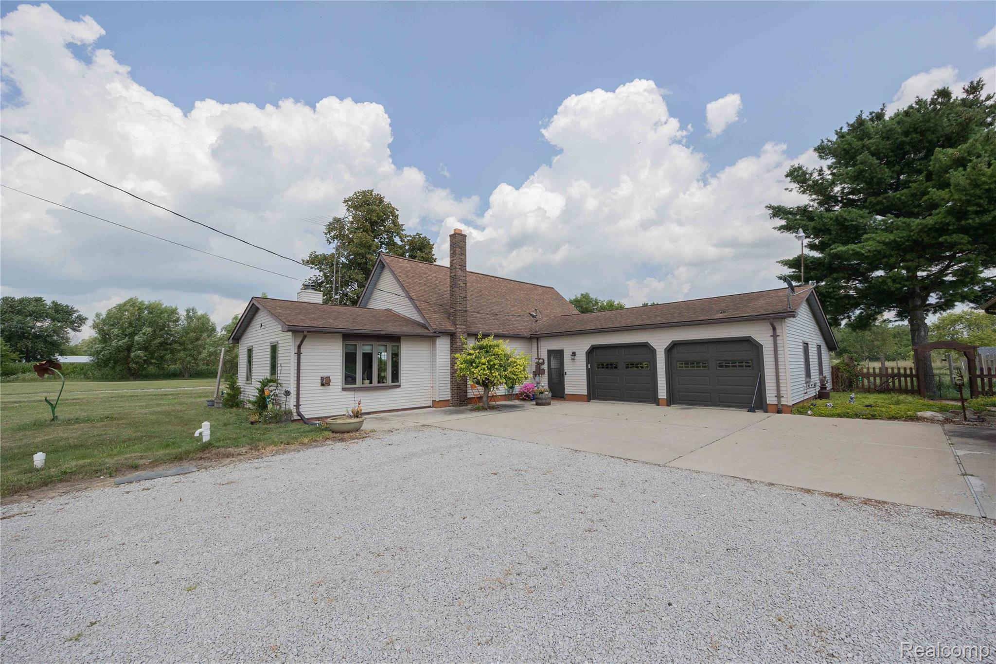 9160 Steffas Road Maybee, MI 48159 - Photo 4 of 36 Expansive gravel driveway, concrete parking pad, attached two-car garage with arched doors, and a brick chimney