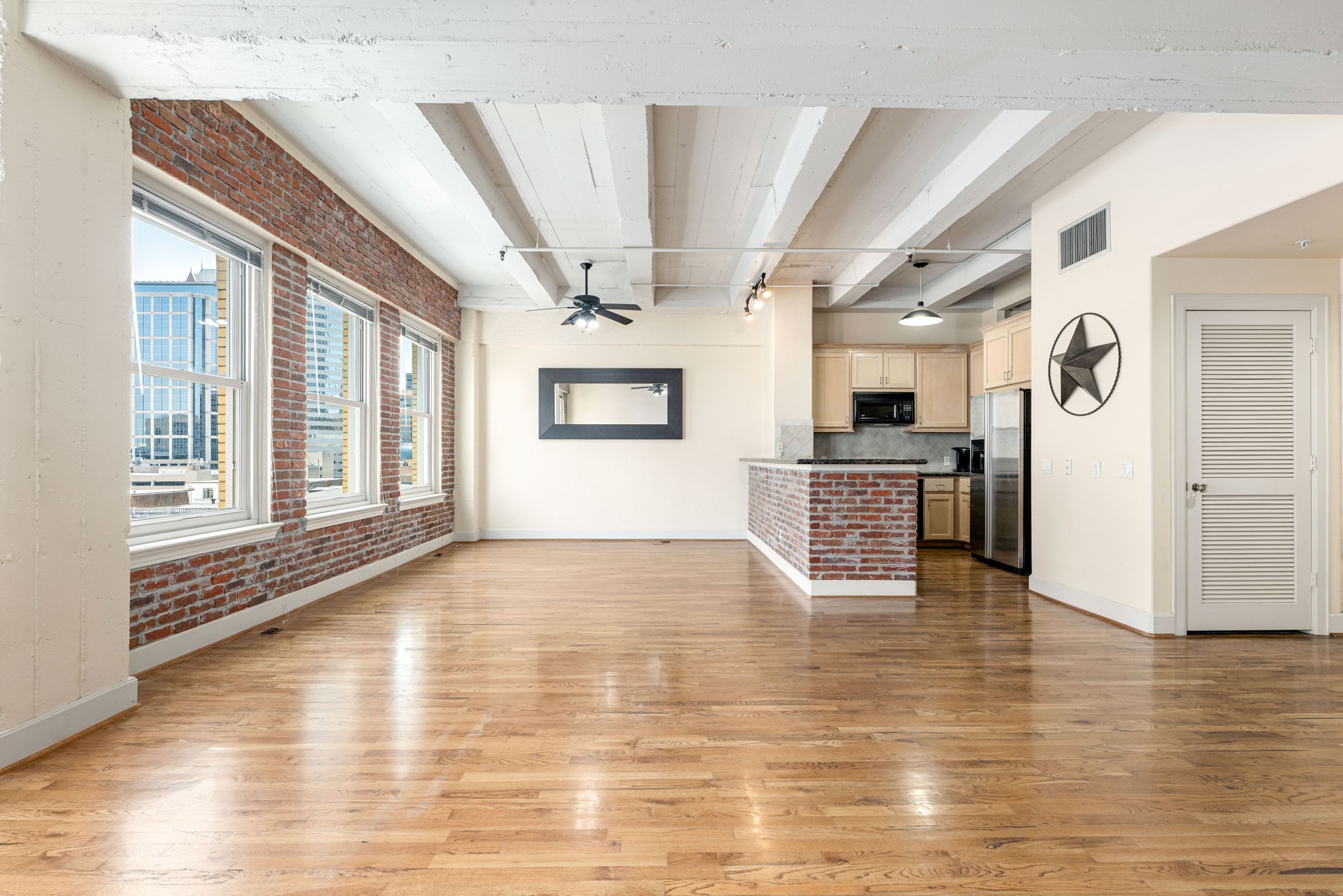 Spacious open-concept living area with warm hardwood floors, exposed brick walls, and abundant natural light from large windows overlooking the city.