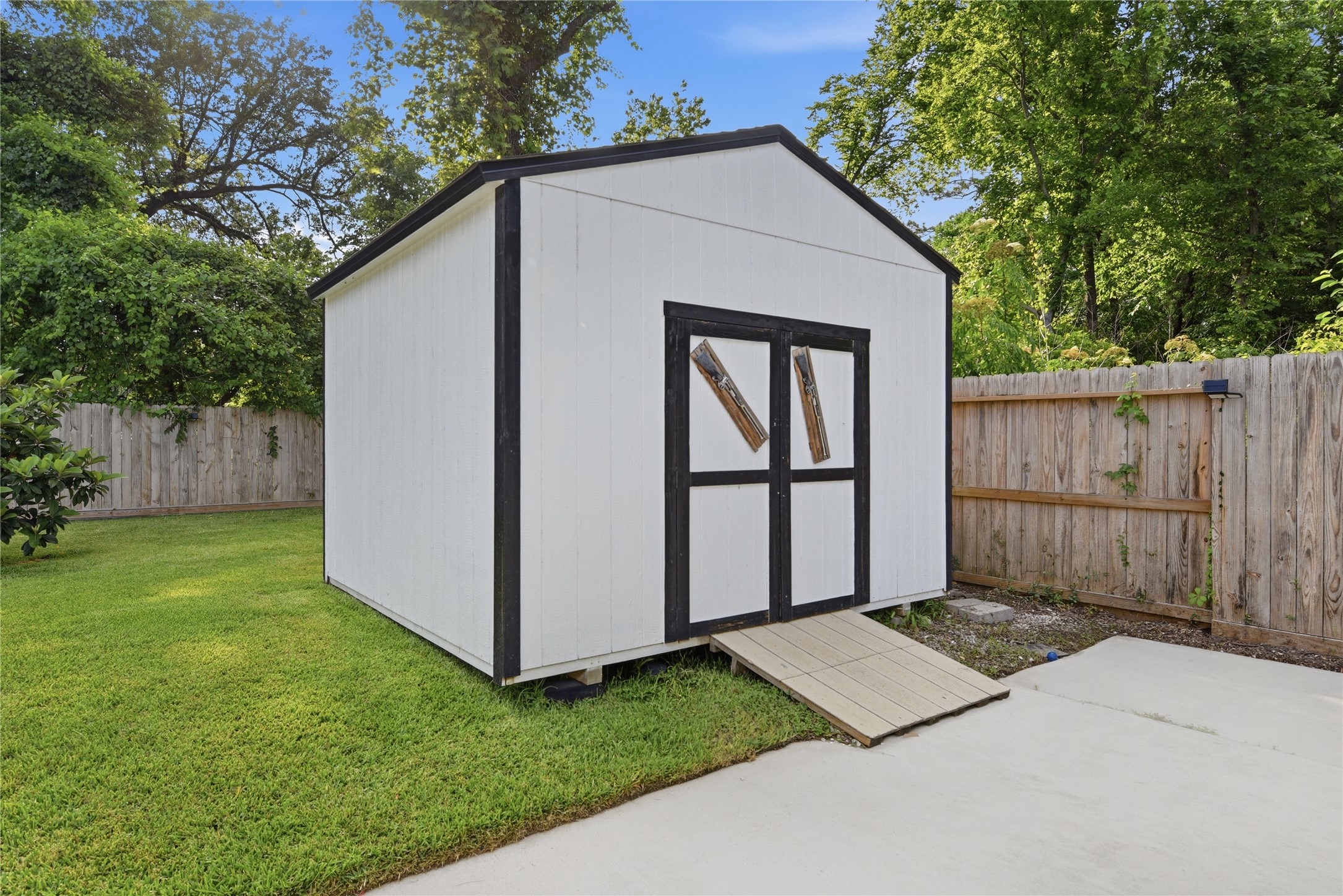 9403 Spaulding Street Houston, TX 77016 - Photo 47 of 50 This photo features a well-maintained white storage shed with black trim in a fenced backyard. It sits on a patch of green lawn, accessible by a small ramp, with lush trees providing a serene backdrop.