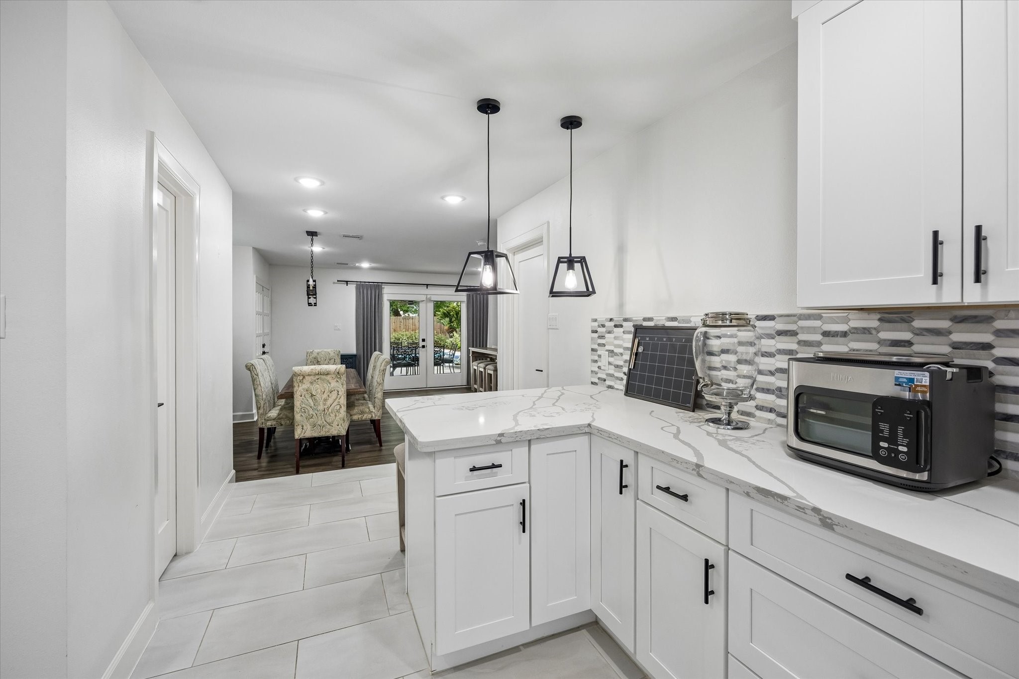 1420 Indiana Street Houston, TX 77006 - Photo 14 of 31 This photo showcases a modern kitchen and dining area with white cabinetry, sleek countertops, and a stylish backsplash. It features pendant lighting and flows into a dining space leading to a patio through glass doors.