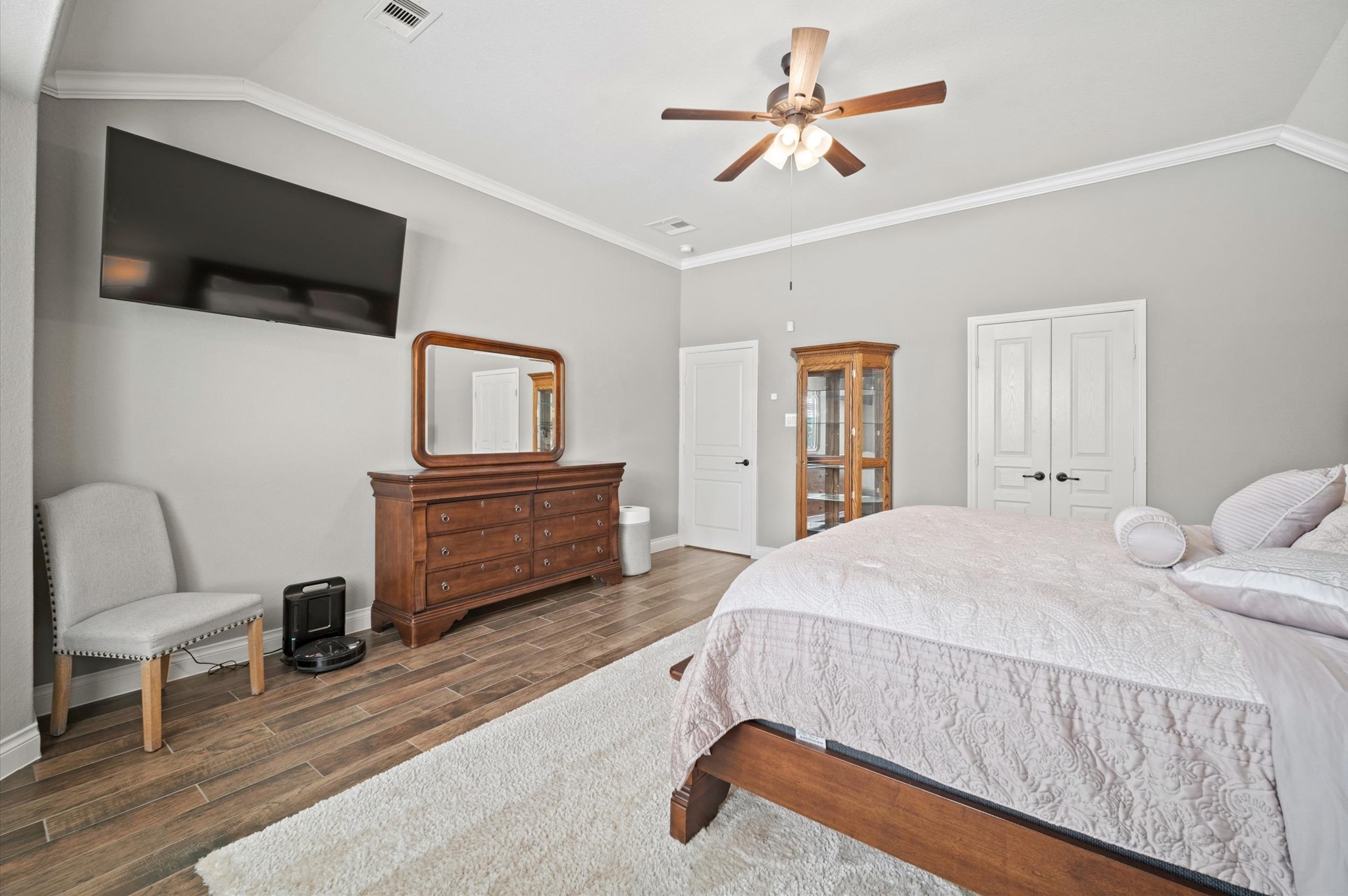 9507 Empress Crossing Drive Spring, TX 77379 - Photo 24 of 50 This spacious bedroom features a stylish ceiling fan, wall-mounted TV, and elegant wood flooring. It includes a large bed, a dresser with a mirror, and ample natural light. Dual doors and tasteful decor provide a cozy yet modern feel.