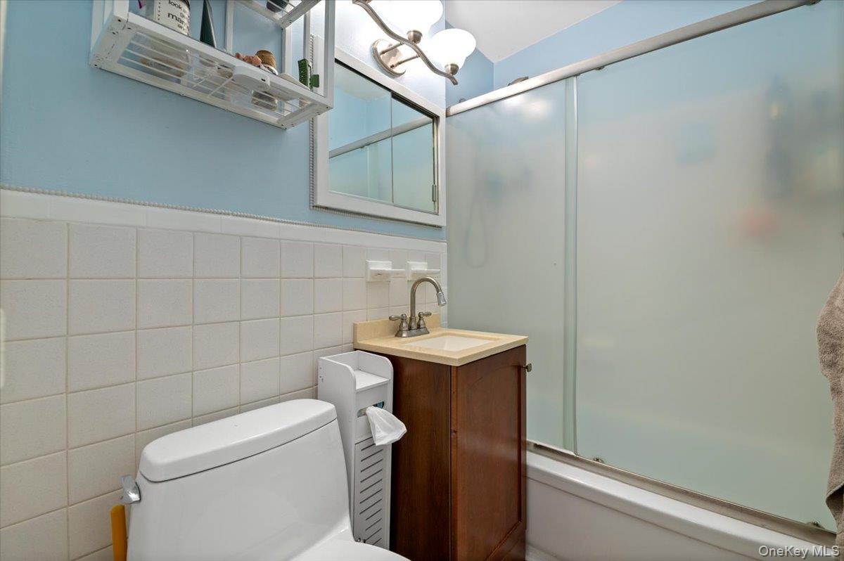 267 North Newbridge Road, Unit 2D Levittown, NY 11756 - Photo 13 of 22 Bathroom featuring white square wall tiling, a built-in tub with a sliding frosted glass enclosure, a wood-finish vanity with an undermount sink, and a wall-mounted light fixture