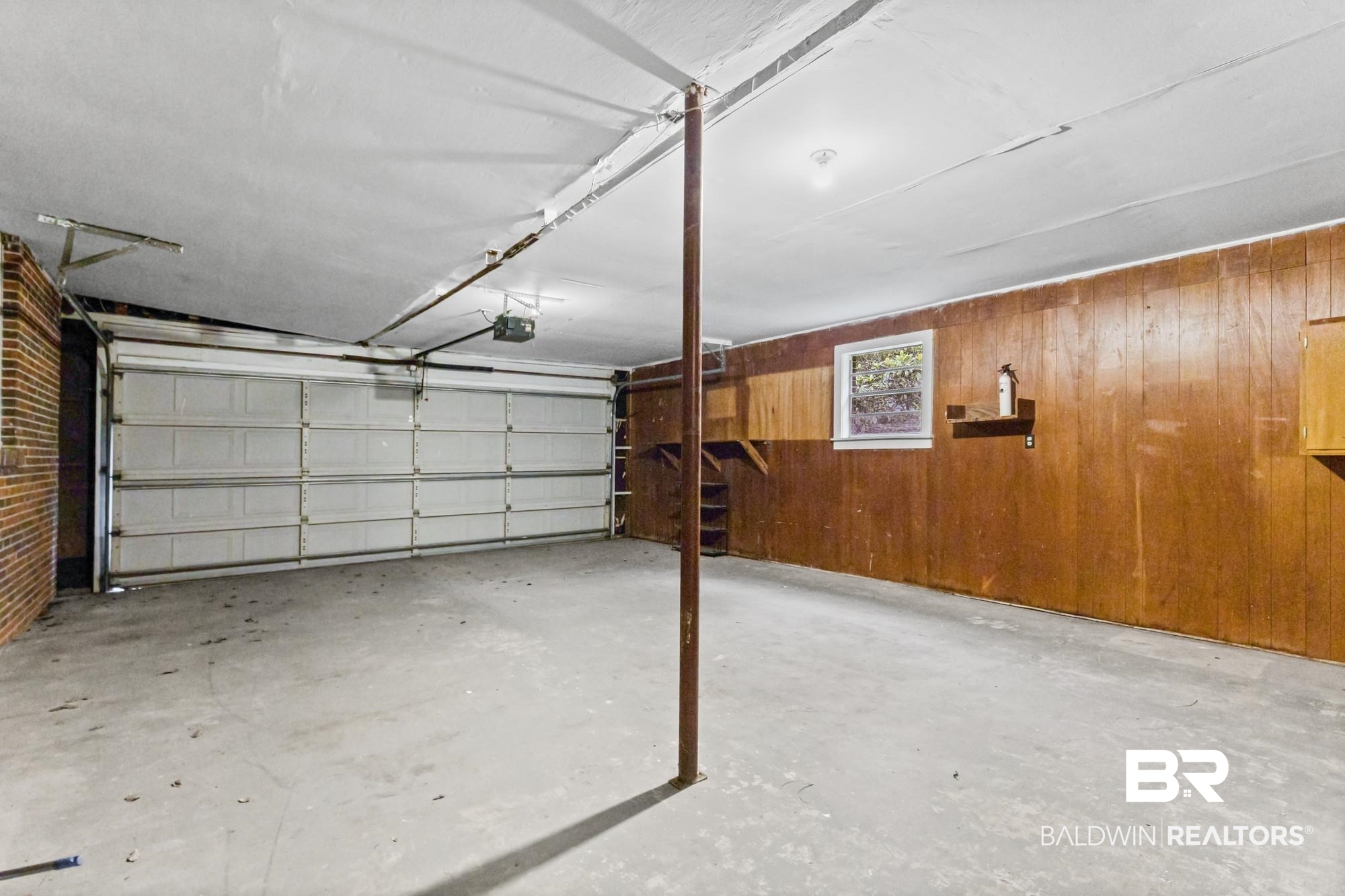 312 Sutherland Drive Mobile, AL 36611 - Photo 24 of 28 Spacious garage featuring a concrete floor, white sectional garage door, wood-paneled walls, a single window, and exposed ceiling joists