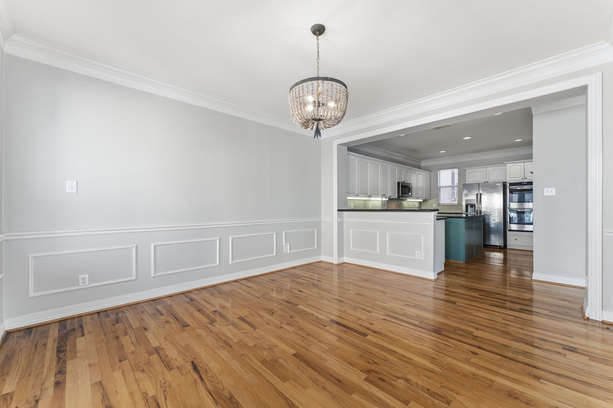 827 Rosine Street Houston, TX 77019 - Photo 27 of 50 This photo showcases a bright, open dining area with elegant wainscoting and hardwood floors, leading into a modern kitchen with white cabinetry and stainless steel appliances. A stylish light fixture adds a touch of sophistication.