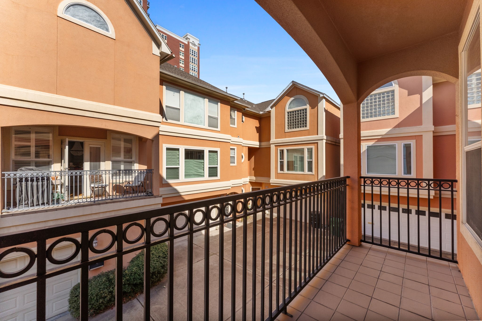 827 Rosine Street Houston, TX 77019 - Photo 28 of 50 This photo features a view from a cozy balcony overlooking a courtyard of a multi-story townhouse complex. The buildings have a warm, peach-colored exterior with large windows, decorative railings, and arched architectural details. The area is well-maintained and offers a charming, community feel.