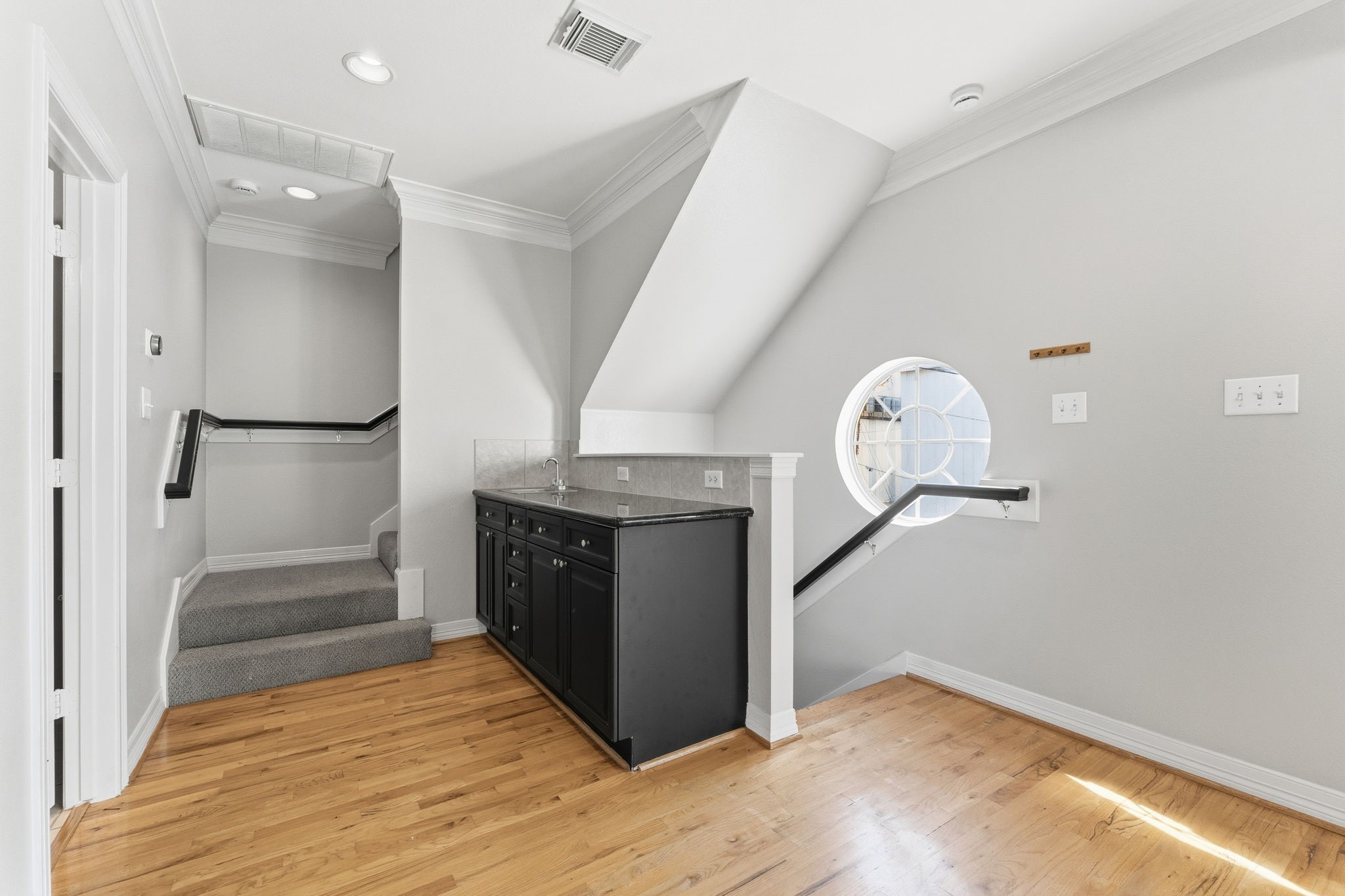 827 Rosine Street Houston, TX 77019 - Photo 30 of 50 This photo shows a bright, modern landing area with hardwood floors and a carpeted staircase. It features a small bar with a sink, black cabinetry, and a unique round window providing natural light. The space is accented by white walls and crown molding, offering a clean and airy feel.