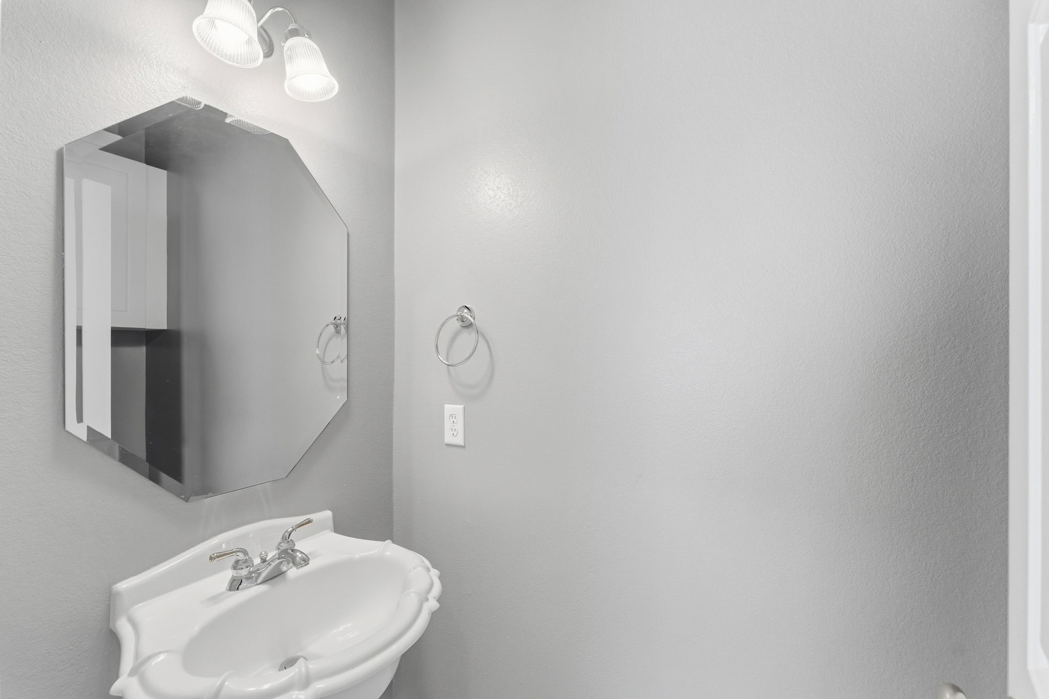 827 Rosine Street Houston, TX 77019 - Photo 33 of 50 This photo shows a small bathroom with a modern, minimalist design. It features a white pedestal sink, an octagonal mirror, and a wall-mounted light fixture above. The walls are painted in a soft gray tone, giving the space a clean and contemporary feel.