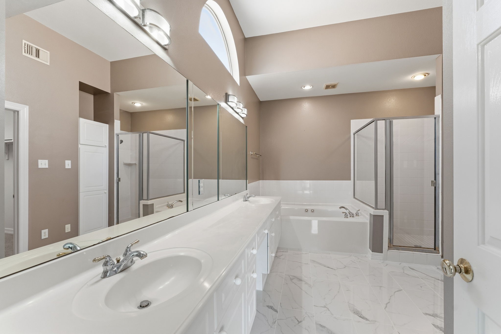 827 Rosine Street Houston, TX 77019 - Photo 39 of 50 This spacious bathroom features a large vanity with dual sinks, expansive mirrors, a soaking tub, and a separate shower. The high ceilings and arched window enhance the bright, airy feel, complemented by elegant tile flooring.