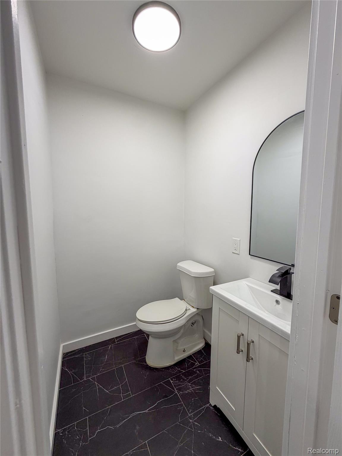 18217 Prairie Street Detroit, MI 48221 - Photo 12 of 34 Compact powder room featuring a white single-sink vanity with matte black faucet, an arched mirror, a toilet, and black marble-look tile flooring