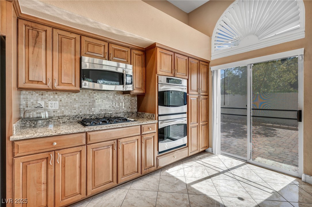 2107 Eaglepath Circle Henderson, NV 89074 - Photo 33 of 84 Kitchen featuring backsplash, light stone countertops, brown cabinets, stainless steel appliances, and light tile patterned flooring