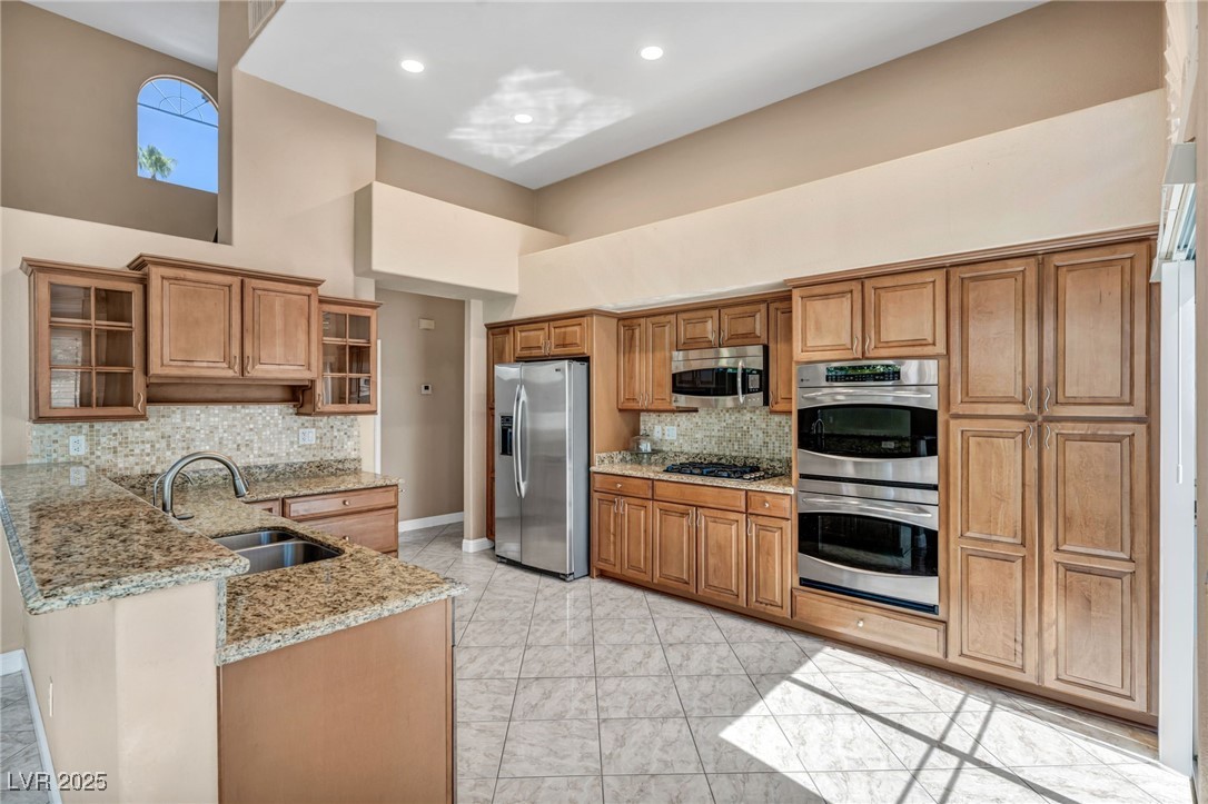2107 Eaglepath Circle Henderson, NV 89074 - Photo 50 of 84 Kitchen featuring glass insert cabinets, light stone counters, brown cabinetry, a towering ceiling, and stainless steel appliances
