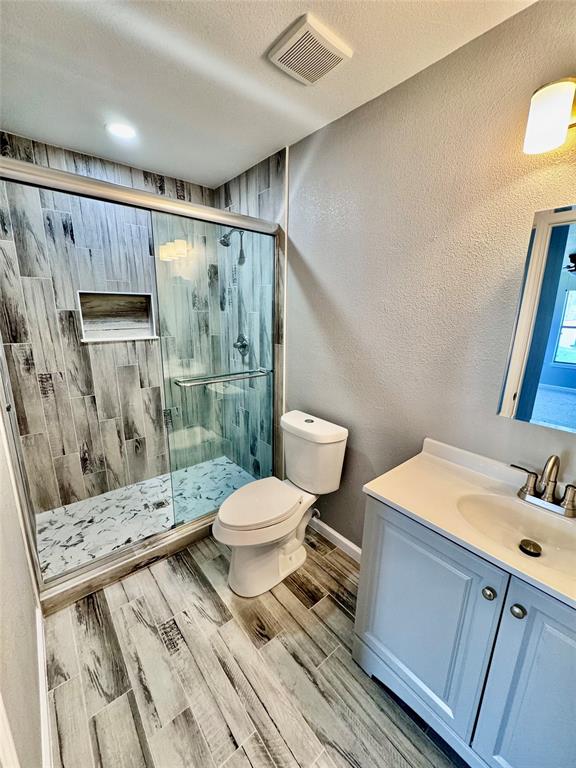 1736 Ransom Terrace Fort Worth, TX 76112 - Photo 16 of 27 Main ensuite Bathroom featuring a glass-enclosed shower with wood-finish tile surround and integrated niche, a white toilet, a vanity with a white countertop and basin sink, and wood-finish tile flooring