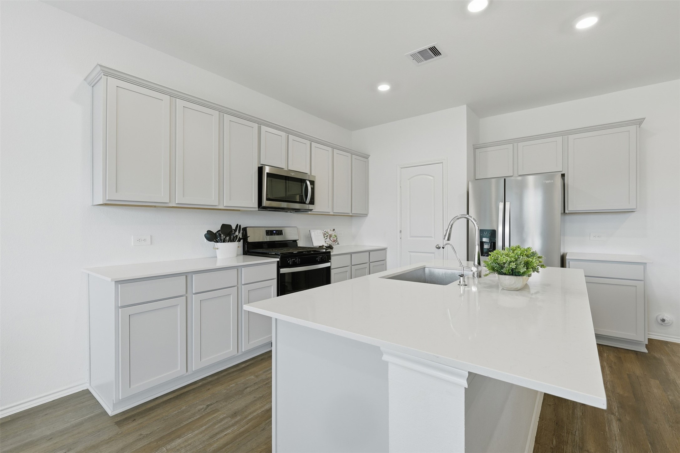 17719 Winfall Drive Crosby, TX 77532 - Photo 13 of 47 Modern kitchen with sleek white cabinetry and countertops, stainless steel appliances, and a spacious island with a sink. The room is bright with recessed lighting and wood flooring.