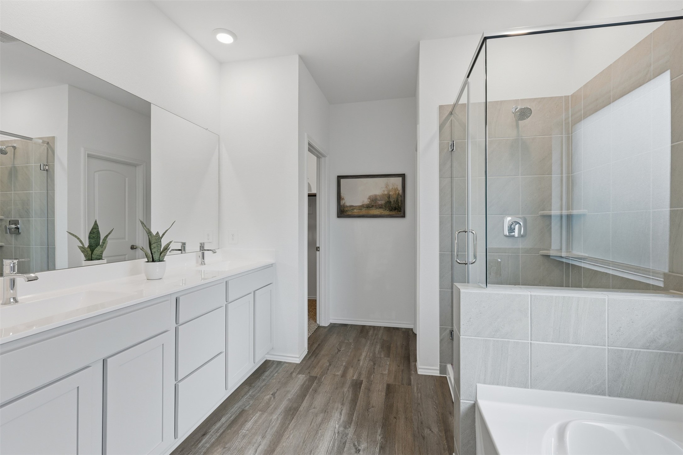 17719 Winfall Drive Crosby, TX 77532 - Photo 20 of 47 This primary bathroom features a modern design with a spacious double vanity, sleek white cabinetry, and a large mirror. The glass-enclosed shower has stylish tile work, and the room is accented by wood-look flooring and neutral tones, creating a bright and inviting space.