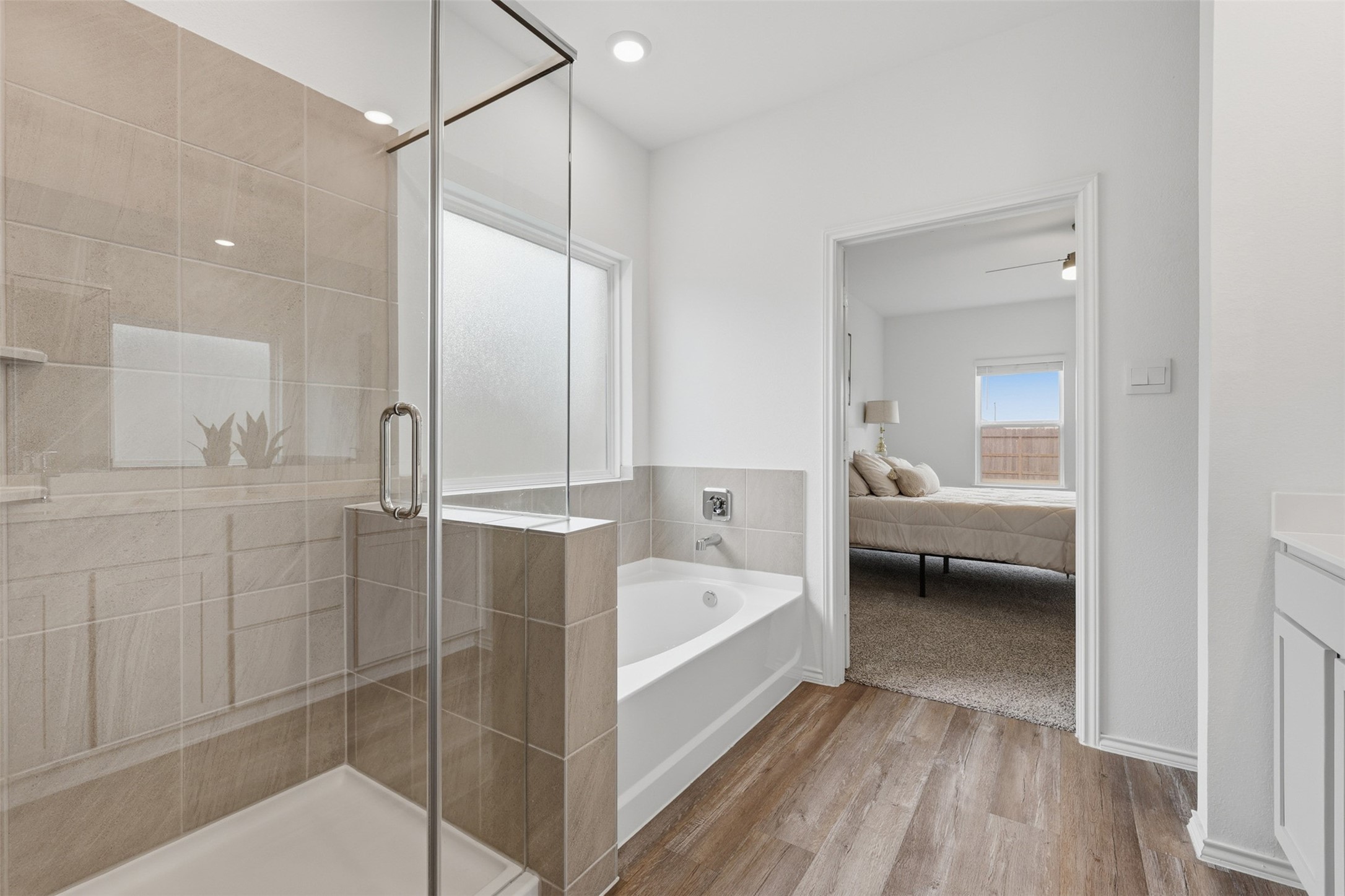 17719 Winfall Drive Crosby, TX 77532 - Photo 21 of 47 This modern primary bathroom features a spacious glass-enclosed shower and a separate bathtub, with sleek tile work and wood-style flooring. It leads into a cozy bedroom with carpeted flooring and a large window for natural light.