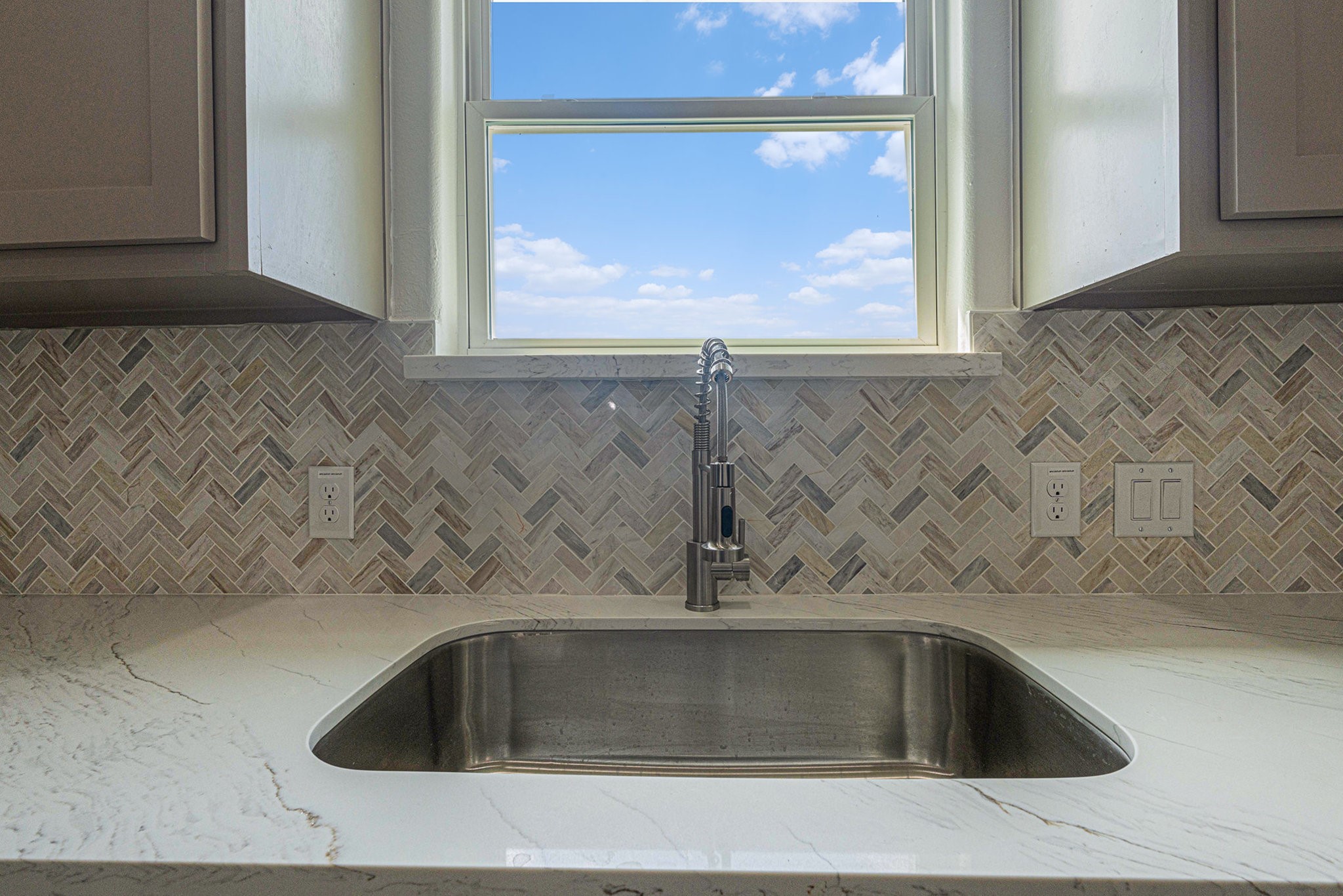 702 Azalea Street Waller, TX 77484 - Photo 33 of 45 This kitchen features a sleek, modern sink with a stainless steel faucet, set in a marble countertop. The stylish herringbone-patterned backsplash adds elegance, while the window above the sink offers natural light and a view.