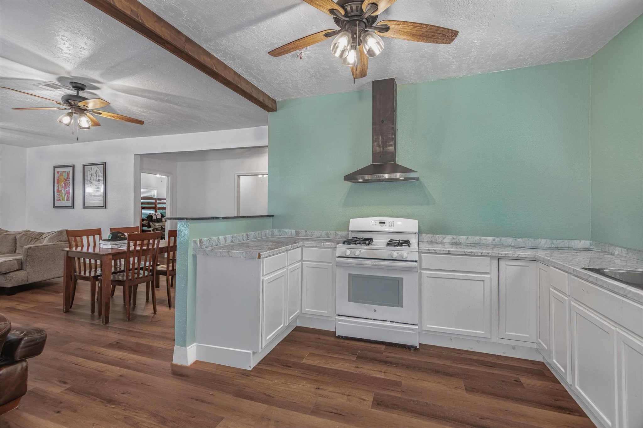 905 43rd Street Galveston, TX 77550 - Photo 35 of 43 This photo features an open-concept kitchen and dining area with hardwood floors. The kitchen has white cabinets, a marble countertop, and a gas stove with a modern hood. The space is brightened by ceiling fans with lights, and there's a cozy living area visible in the background.