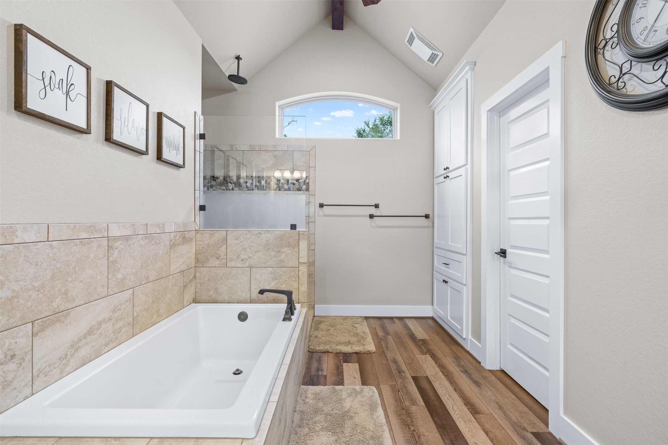 1306 22 Hills Road Gause, TX 77857 - Photo 4 of 40 Bathroom featuring a built-in soaking tub with dark bronze fixtures, a walk-in shower with glass enclosure and tiled walls, wood-finish flooring, a vaulted ceiling, and an arched window