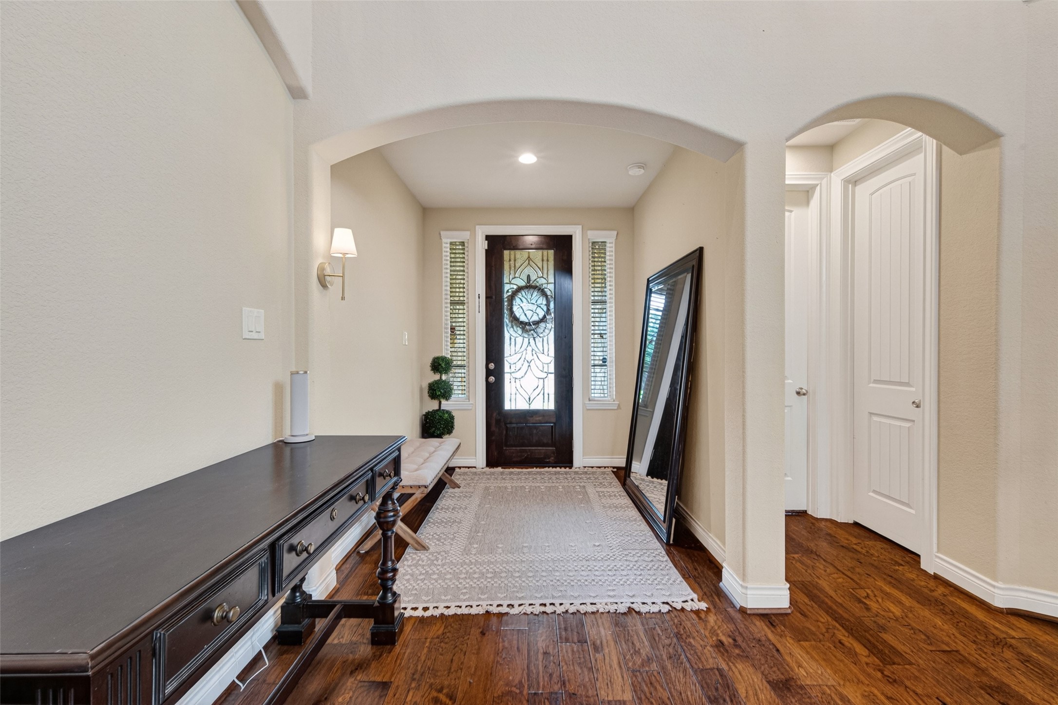 25611 Ellis Ridge Court Katy, TX 77494 - Photo 5 of 49 This inviting entryway features a stylish dark wood door with decorative glass, warm hardwood floors, and arched doorways. The space is accented by a side table, wall sconce, and a large mirror, creating a welcoming atmosphere.