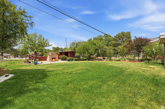 7576 South Jefferson Street Midvale, UT 84047 - Photo 39 of 53 Expansive green lawn with a stone outdoor fireplace feature, a wood-decked area, a red storage shed, and mature trees outlining the property