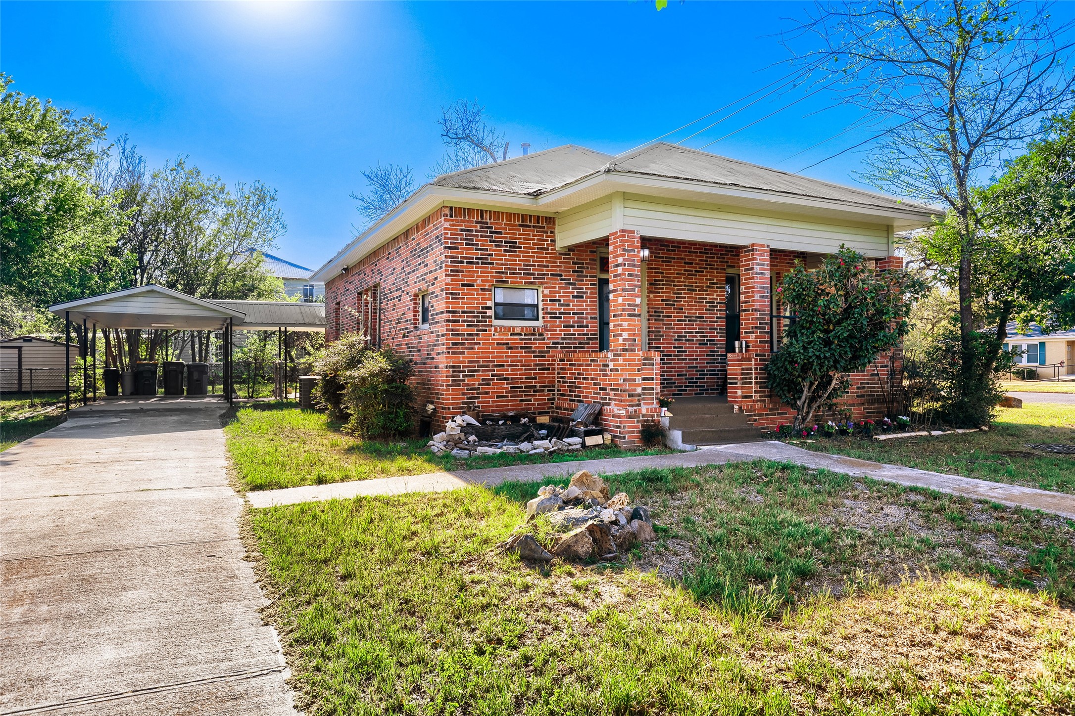 Charming brick home with a classic porch, surrounded by greenery. It features a spacious driveway leading to a covered carport, with mature trees adding privacy and shade. Perfect for those seeking a cozy and inviting exterior.