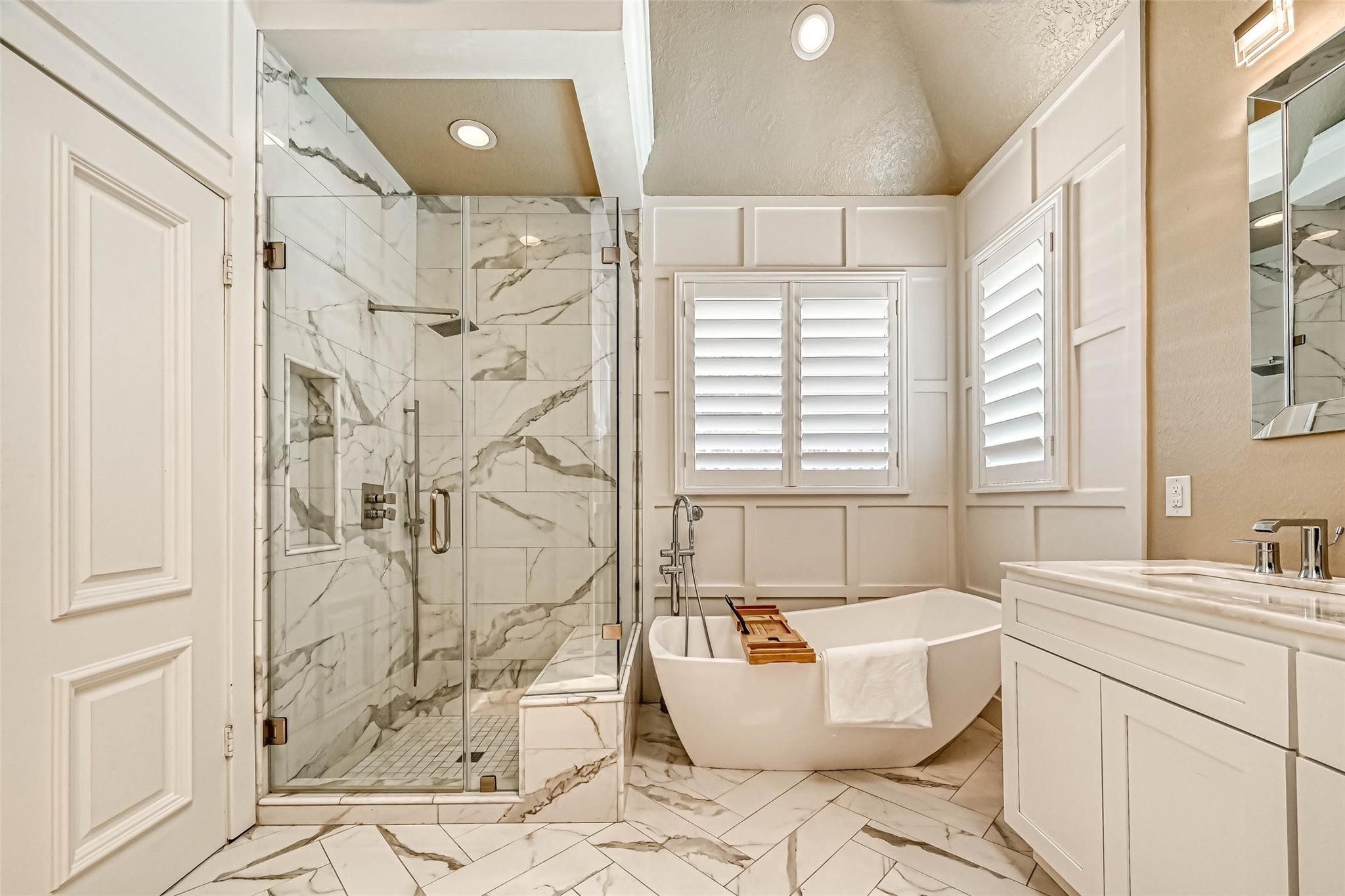 263 Wood Loop Street Houston, TX 77015 - Photo 5 of 45 This bathroom features a modern design with a spacious glass-enclosed shower and a freestanding bathtub. It has elegant white marble tiles, a double-sink vanity, and natural light streaming through shuttered windows, creating a bright and luxurious atmosphere.