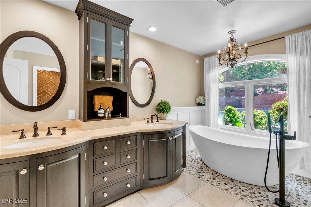 108 Quail Run Road Henderson, NV 89014 - Photo 35 of 50 Full bath with double vanity, a freestanding bath, a chandelier, light tile patterned floors, and wainscoting