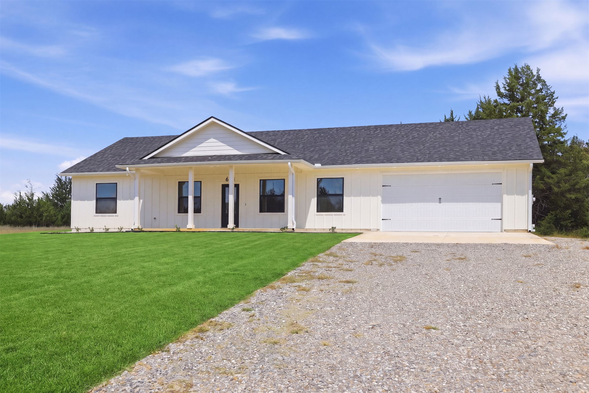 63 Deerfield Road Colbert, OK 74733 - Photo 2 of 33 Single-story residence featuring white board-and-batten siding, a dark shingle roof, a covered front porch with white columns, dark-framed windows, and an attached two-car garage with a white door