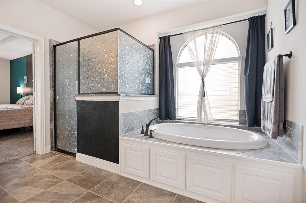 413 Frontier Court Midlothian, TX 76065 - Photo 17 of 26 Primary bathroom with soaking tub set against an arched window, complemented by a textured glass shower enclosure with a dark frame, and finished with large format tile flooring and wainscoting