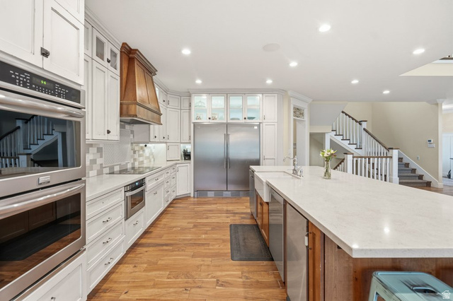 672 Perry Hollow Drive Mapleton, UT 84664 - Photo 29 of 106 Gourmet kitchen featuring a large central island with a farmhouse sink, stainless steel appliances, wood-finish flooring, white cabinetry, and a custom wood range hood