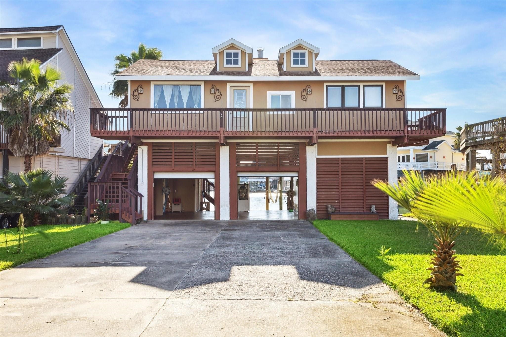 This charming waterfront home features a raised design with a large wooden balcony, lush green lawn, and space underneath for parking or storage. Its inviting facade includes dormer windows and decorative accents, perfect for enjoying serene canal views.