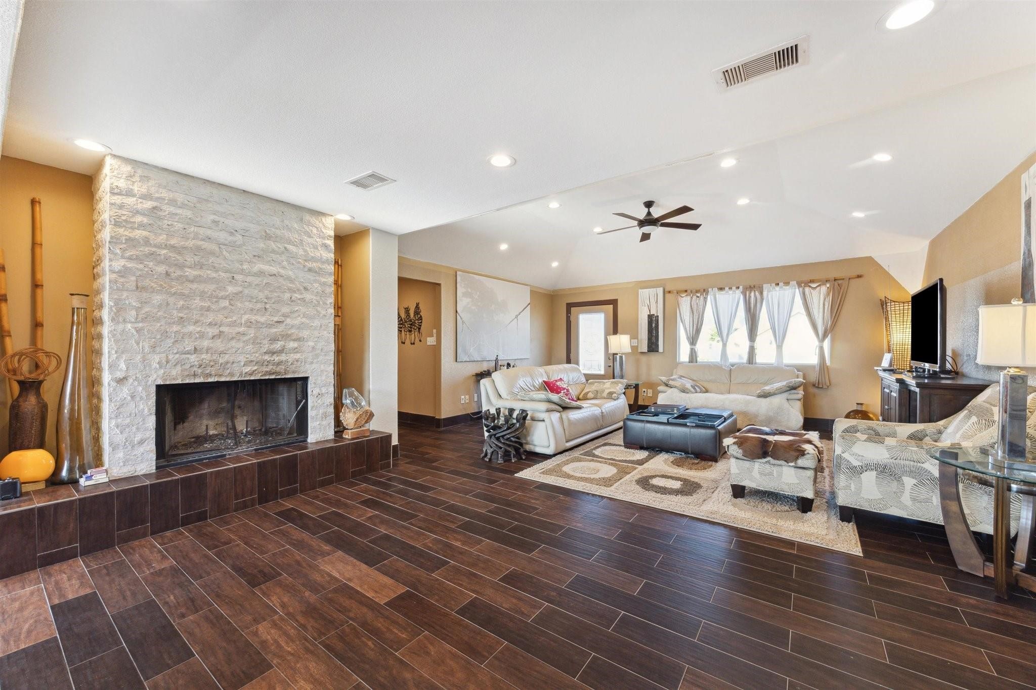 410 Isles End Road Tiki Island, TX 77554 - Photo 2 of 13 This spacious living room features a cozy stone fireplace, dark wood-look tile flooring, and vaulted ceilings with recessed lighting. It's elegantly furnished with comfortable seating, a large TV, and plenty of natural light from the curtained windows.